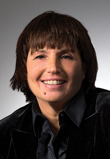 A person with short dark hair and a mole on their cheek smiles at the camera. They are wearing a dark jacket over a collared shirt. The background is a neutral gray.