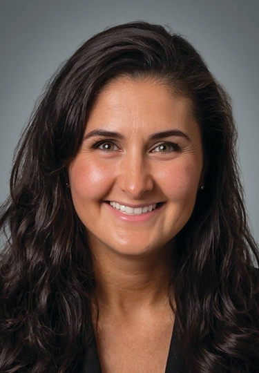 A woman with long, wavy dark hair smiles at the camera. She is wearing a black top and has a light complexion. The background is a plain, neutral gray.