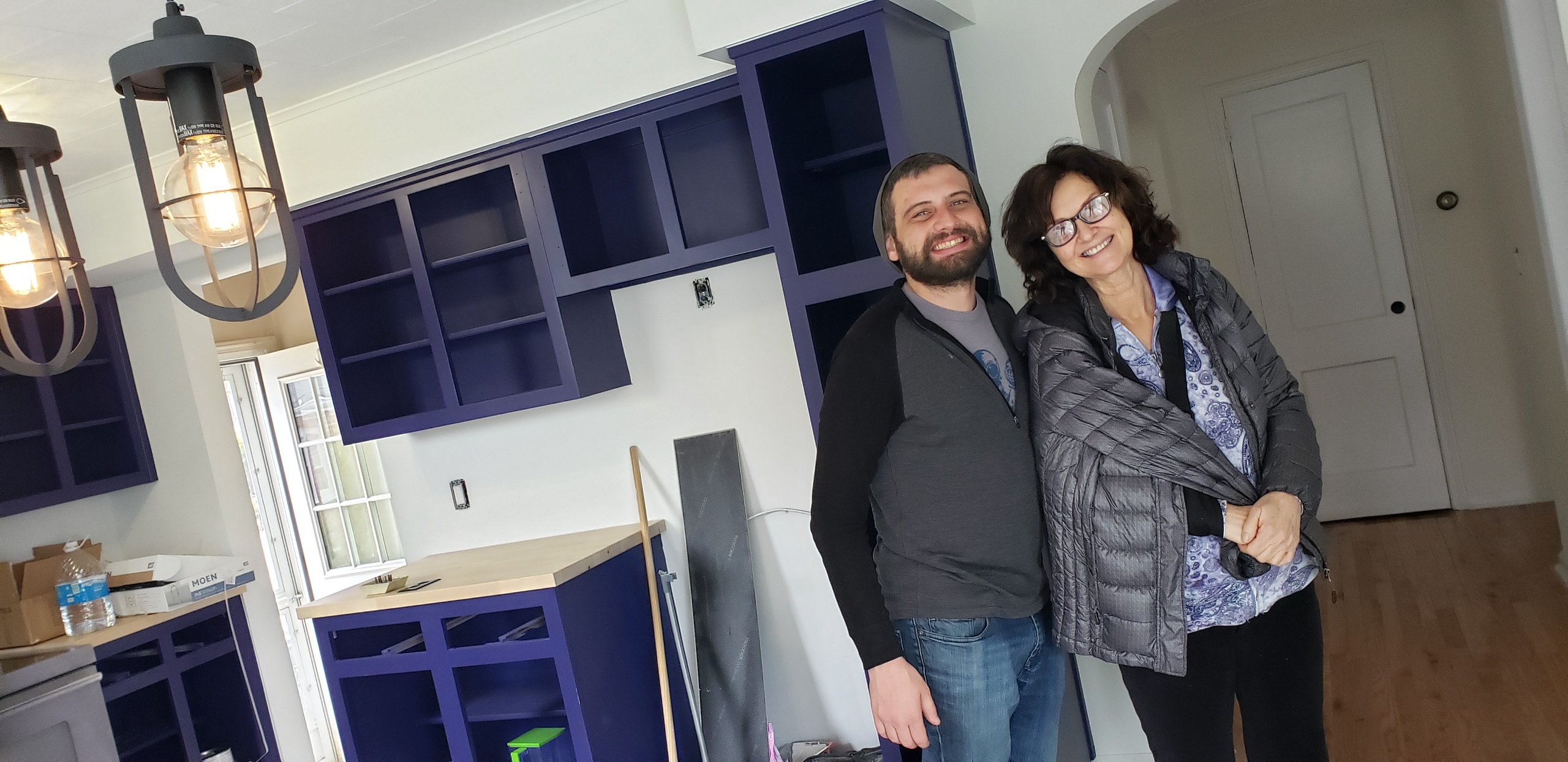 Two people stand smiling in a room with unfinished purple cabinets. The room has a modern light fixture and wooden floor. The man wears a long-sleeve shirt, and the woman wears glasses and a jacket.