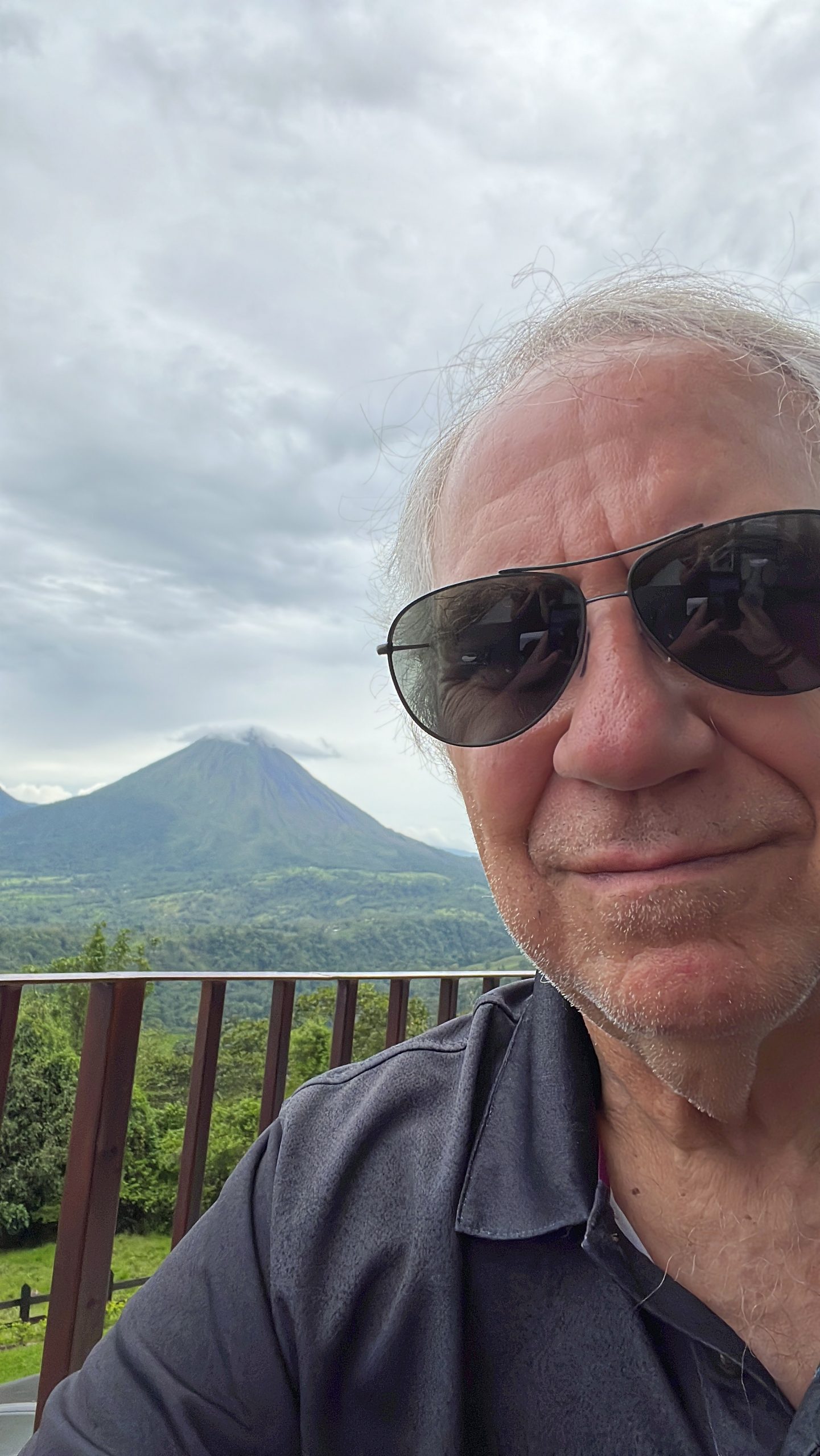 A person with sunglasses takes a selfie on a balcony with a scenic view of a distant volcano under a cloudy sky.