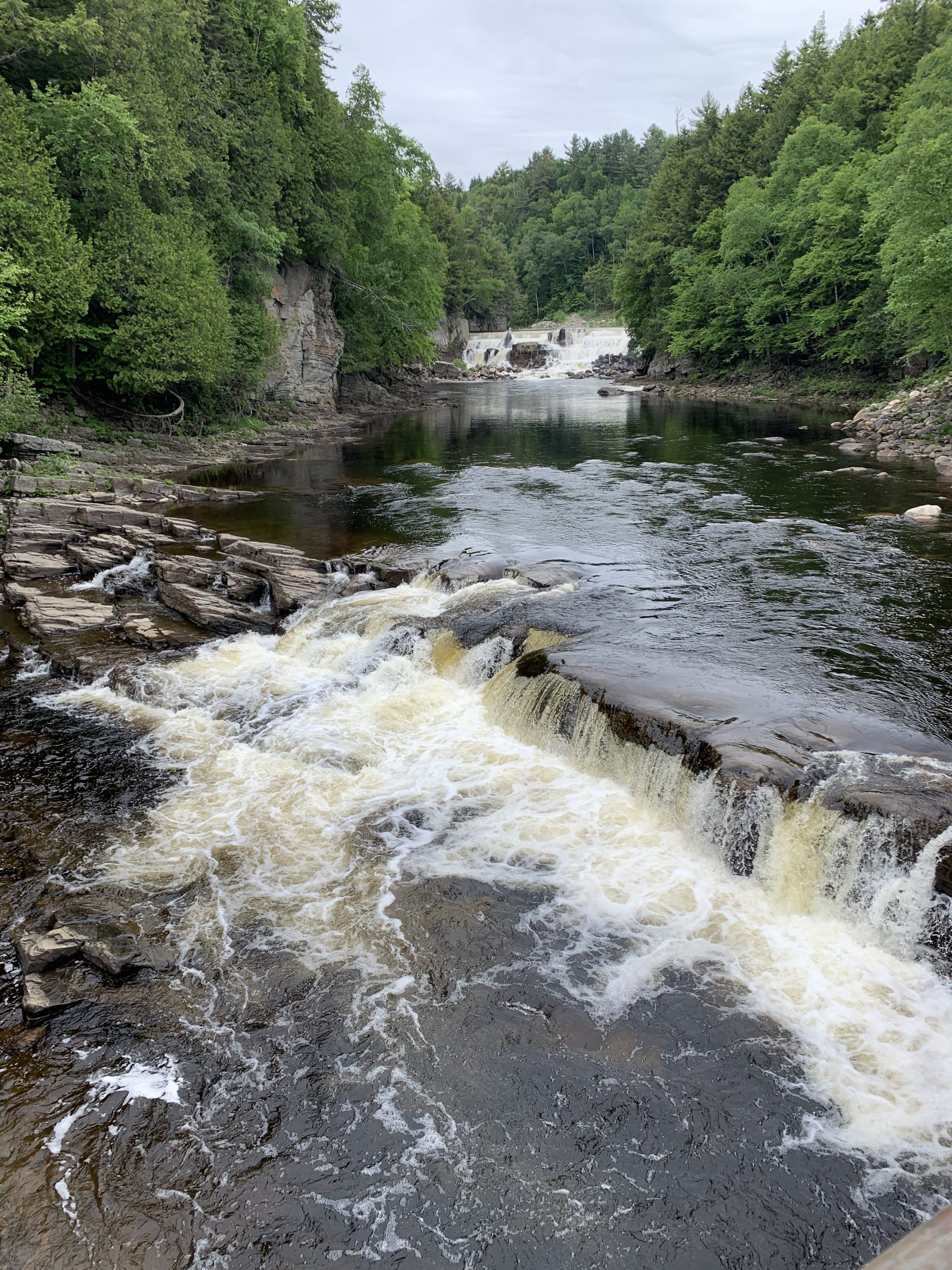 A scenic view of a river flowing over rocks surrounded by lush green trees. In the background, a waterfall cascades down, set against a backdrop of dense forest under an overcast sky.