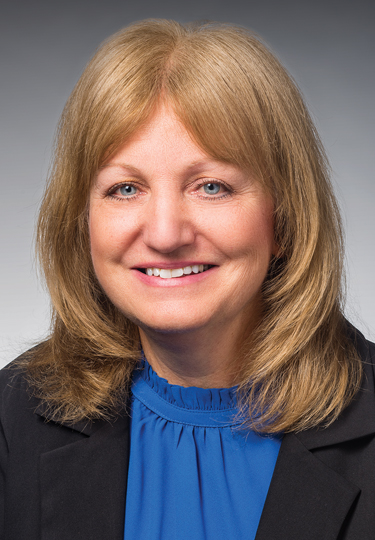A woman with shoulder-length blonde hair is smiling. She is wearing a blue blouse and a dark blazer. The background is a neutral gradient. headshot