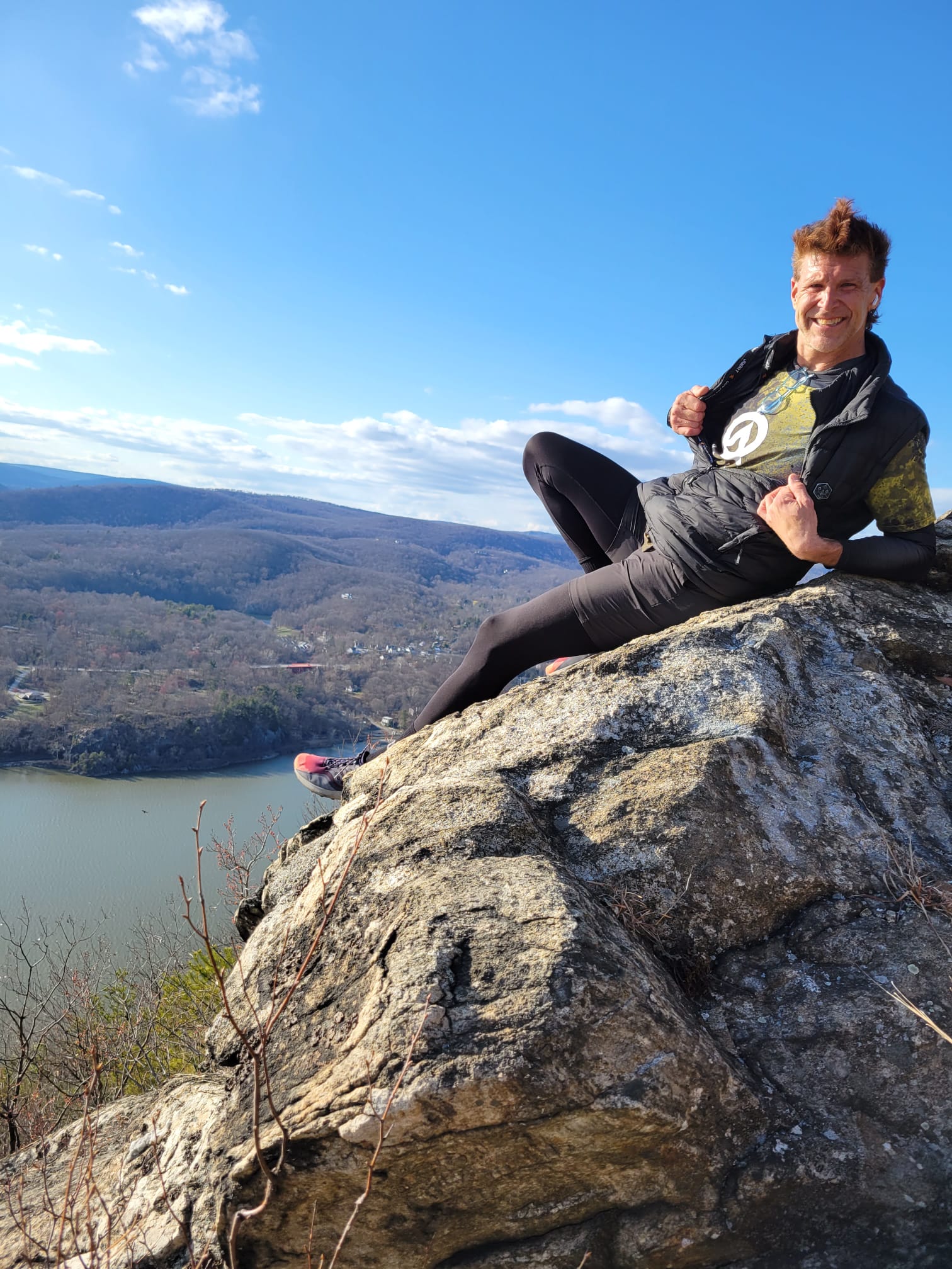 A person in athletic gear poses confidently on a rocky cliff overlooking a vast landscape with a river and distant hills under a bright blue sky.