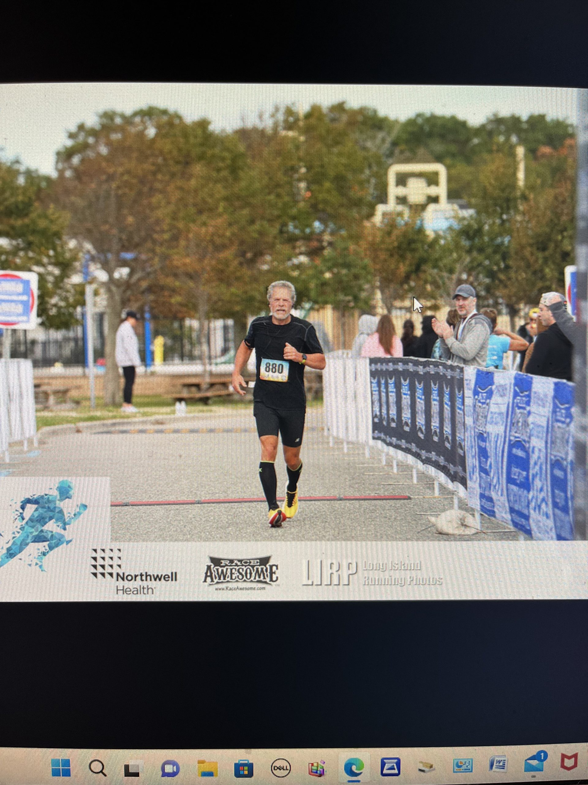A man wearing a black athletic outfit and race bib number 880 runs on a race track. Spectators are visible on either side, standing behind barriers. Trees and a blue sky are in the background.