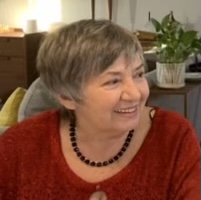 A woman with short gray hair smiles while wearing a red sweater and a black beaded necklace. She is indoors, with a plant and furniture visible in the background.