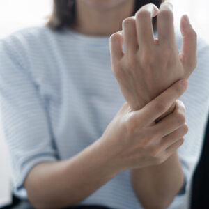 A person wearing a light blue and white striped shirt holds their right wrist with their left hand. The image focuses on the hands and wrists, suggesting possible wrist pain or discomfort.