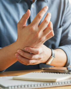 A person wearing a blue shirt is sitting at a table, massaging their left hand with their right. A smartphone and notebooks are on the table in front of them.