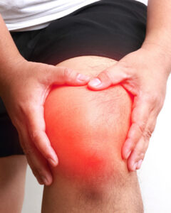 A person wearing black shorts holds their knee with both hands. The knee area is highlighted with a red glow, suggesting pain or inflammation.