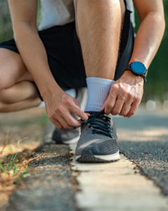 A person in black shorts kneels on a road, tying the laces of a gray running shoe. They wear a white sock and a smart wristwatch. Sunlight highlights the scene, with trees and greenery in the blurred background.