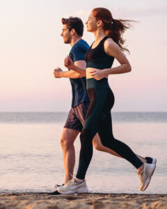 A man and woman jog side by side along a beach at sunset, wearing athletic clothing and sneakers, with the calm sea and pastel sky in the background.