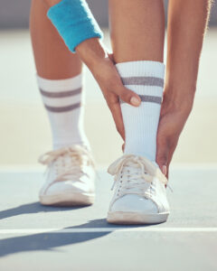 A person wearing white sneakers, white socks with gray stripes, and a blue wristband bends down to touch their ankle on a sports court.