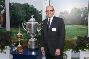 A man in a suit and glasses stands indoors next to two large golf trophies on a table, with a golf course scene displayed in the background.
