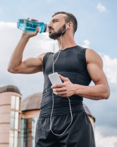 A fit man in a black sleeveless shirt drinks water from a blue bottle while holding a smartphone and wearing wired earphones outdoors, with modern buildings and a blue sky in the background.