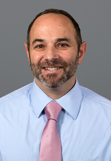 A man with short dark hair and a beard smiles at the camera. He is wearing a light blue shirt and a light pink tie, set against a plain gray background. headshot