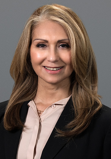 A woman with long, light brown hair and subtle highlights smiles at the camera. She is wearing a light beige blouse and a black blazer, posed in front of a plain gray background. headshot