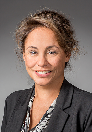 A woman with wavy brown hair pulled back, wearing a black blazer and patterned blouse, smiles at the camera against a plain gray background.