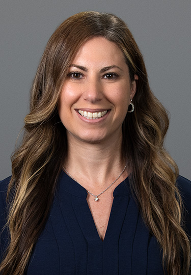 A woman with long, wavy brown hair smiles at the camera. She is wearing a dark blue top, a silver necklace, and hoop earrings. The background is plain and gray. headshot