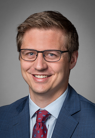 A man with short light brown hair, glasses, and a blue suit with a red patterned tie, smiling in front of a plain gray background. headshot