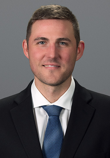 A man with short brown hair wearing a black suit, white shirt, and blue tie, smiling slightly, posed against a plain gray background. headshot