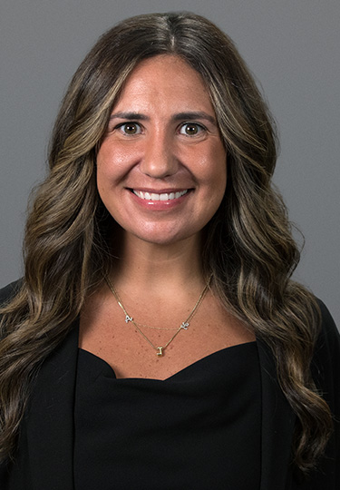 A woman with long, wavy brown hair wearing a black top, black jacket, and a gold necklace with letter charms, smiling in front of a plain gray background. headshot