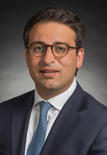 A man wearing glasses, a dark suit, white shirt, and blue patterned tie poses for a professional headshot against a dark, gradient background. headshot