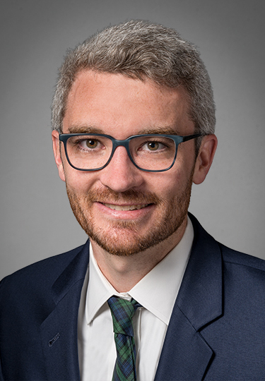 A man with short gray hair, a beard, and glasses is wearing a navy suit, white shirt, and green plaid tie, smiling at the camera against a plain gray background. headshot
