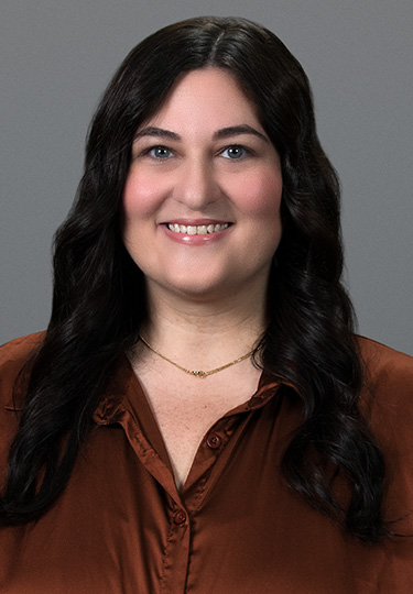 A woman with long, wavy dark hair is smiling at the camera. She is wearing a brown blouse and a delicate gold necklace, posed in front of a plain gray background. headshot