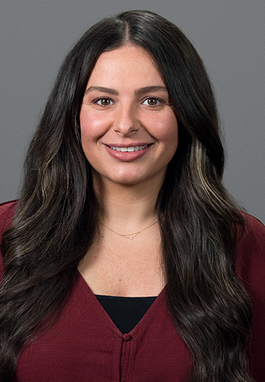 A woman with long, dark wavy hair is smiling at the camera. She is wearing a burgundy top over a black camisole and a delicate necklace. The background is plain and gray. headshot