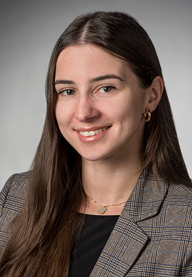 A woman with long brown hair, wearing a plaid blazer, black top, gold hoop earrings, and a necklace, smiling at the camera against a gray background.