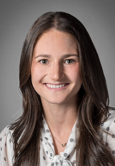 A woman with long brown hair and brown eyes smiles at the camera. She is wearing a white blouse with gray polka dots and a small necklace, set against a plain gray background.