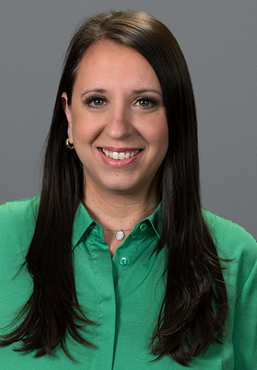A woman with long dark brown hair, wearing a green button-up shirt and small hoop earrings, smiles at the camera against a plain gray background. headshot