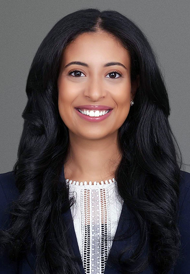 A woman with long, wavy black hair smiles at the camera. She is wearing a dark blazer over a white top with a decorative neckline. The background is plain gray. headshot