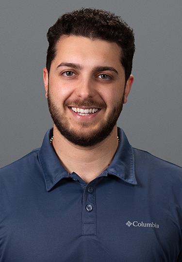 A man with short curly dark hair and a beard smiles at the camera. He is wearing a blue Columbia polo shirt and stands in front of a plain gray background. headshot