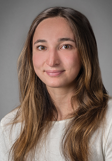 A young woman with long, straight brown hair, wearing a white top, smiles softly at the camera against a plain gray background.