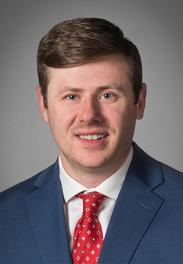 A man with short brown hair in a blue suit, white shirt, and red patterned tie is smiling at the camera against a plain gray background.
