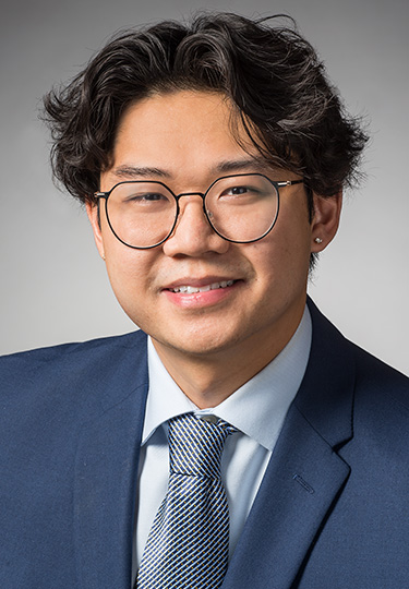 A young man with wavy dark hair, wearing round glasses, a blue suit, light blue shirt, and patterned tie, smiles at the camera against a plain gray background.