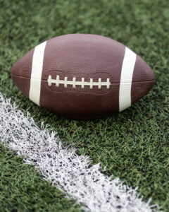 A brown American football with white stripes and laces rests on artificial turf, positioned next to a white line marking on the field.
