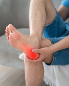 A person sitting on a couch holds their bare foot, with a red area on the heel indicating pain or inflammation. They are wearing denim shorts, and their foot rests on their opposite knee.