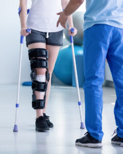 A person wearing a knee brace and using crutches is walking indoors, while a healthcare professional in blue scrubs stands nearby, providing support and guidance.
