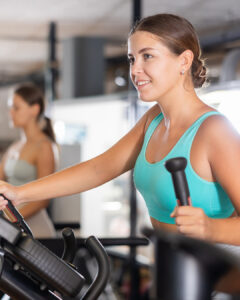 A woman in a turquoise sports bra exercises on an elliptical machine at a gym, smiling and looking ahead. Another woman in workout attire is blurred in the background, also using gym equipment.