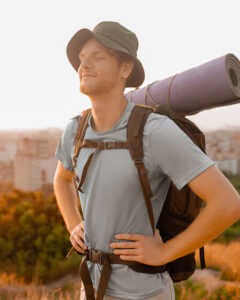 A young man with a backpack and a rolled-up sleeping mat stands outdoors, eyes closed and smiling, enjoying the sunlight. He wears a light shirt and a bucket hat, with a city and greenery in the background.