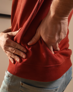 A person wearing a red shirt and blue jeans holds their lower back with both hands, suggesting discomfort or back pain.