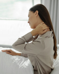 A woman in a beige shirt sits on a bed, touching her neck with a pained expression, suggesting neck discomfort. She is facing sideways with natural light coming through a window in the background.