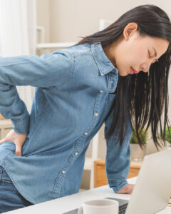 A woman in a denim shirt stands beside a desk, holding her lower back and wincing in pain, suggesting discomfort or back pain, in a bright, home office setting.