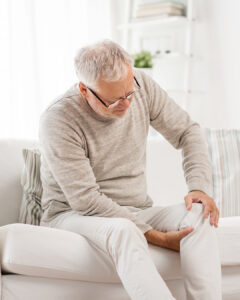An older man with gray hair and glasses sits on a white couch, holding his knee with a pained expression, suggesting he is experiencing knee pain. The background is bright and softly furnished.