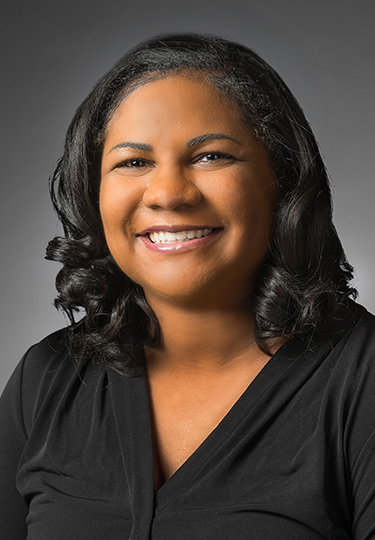 A woman with medium-dark skin and shoulder-length dark curly hair smiles at the camera. She is wearing a black V-neck top and is posed against a neutral gray background.