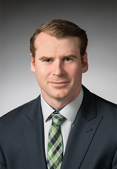 A man with light skin and short, neatly combed brown hair is wearing a dark suit, white shirt with small patterns, and a green plaid tie. He is smiling slightly and posed against a gray gradient background.