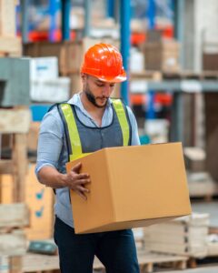 A man wearing a safety vest and orange hard hat carries a large cardboard box in a warehouse filled with shelves and packages.