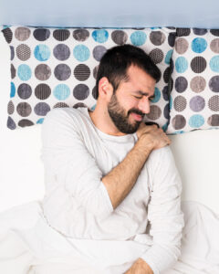 A man with a beard lying in bed, eyes closed, wearing a white shirt, and holding his left shoulder in pain. The bed has pillows with blue and gray polka dot patterns.