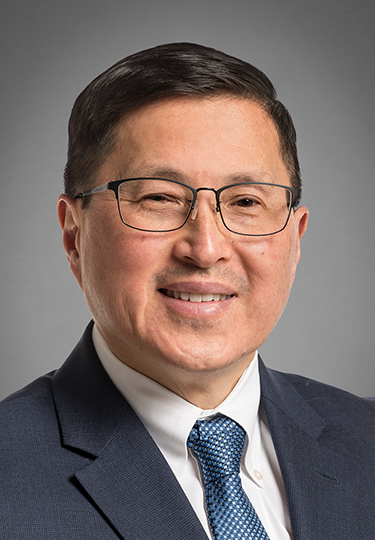 A middle-aged man with short dark hair, glasses, and a slight smile is wearing a navy suit, white shirt, and blue patterned tie. He is posed against a plain gray background. headshot
